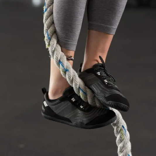 zapatillas running mujer Barefoot negras de malla, agarrándose a una cuerda de entrenamiento, mostrando la flexibilidad y agarre ideal para zapatillas trail running mujer o actividades de cross-training.